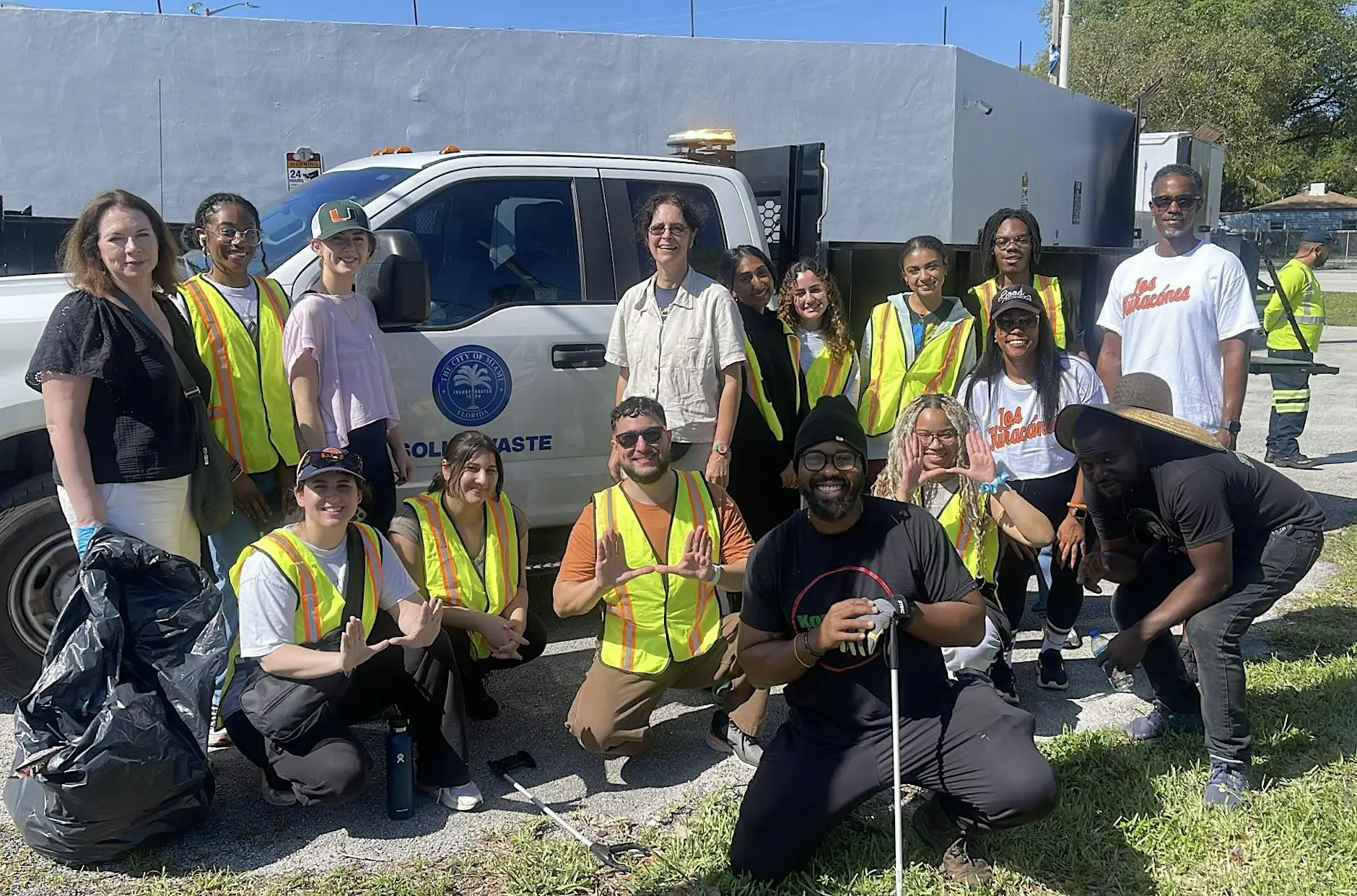 A group of cleanup volunteers in safety vests poses beside a City of Miami solid waste truck with collected trash bags.