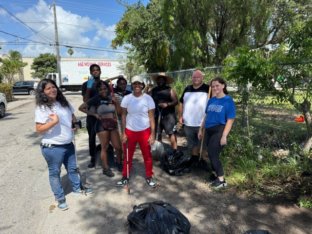 Street cleanup volunteers pose with litter grabbers and filled trash bags beside a tree-lined sidewalk.