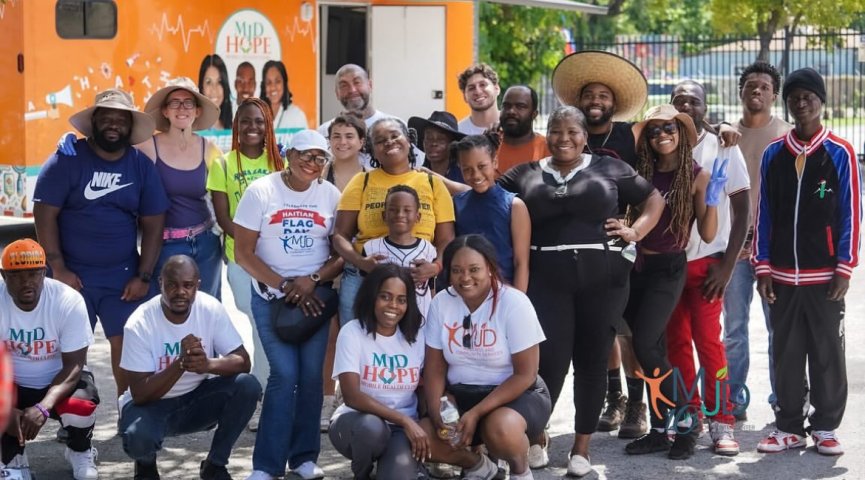 Community members and volunteers gather for a group photo in front of an orange medical outreach vehicle.