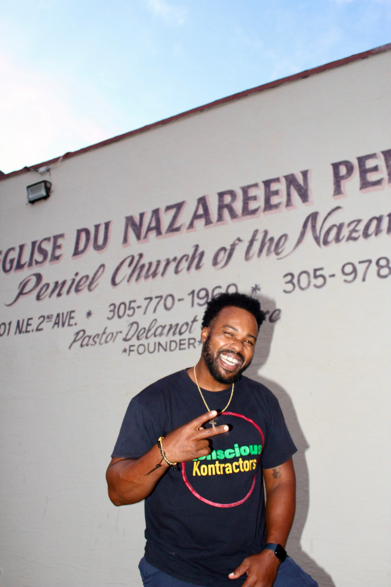 A smiling community member wearing a Konscious Kontractors shirt stands in front of Peniel Church of the Nazarene.