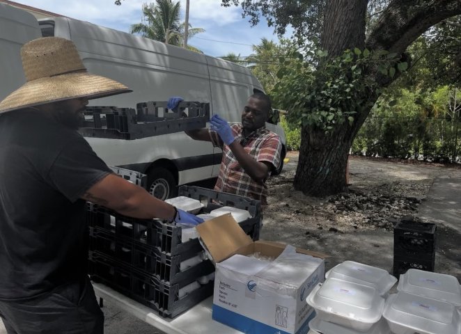 Volunteers wearing gloves organize crates and packaged meals at an outdoor table.